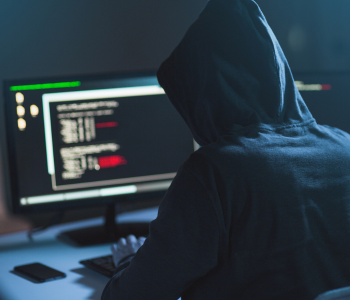 A person in a black hoodie is unidentified as they are typing malicious code on a monitor.