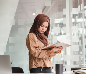 A woman with a headscarf on is reading some paper documents in her hands. A white office with glass windows is the background.
